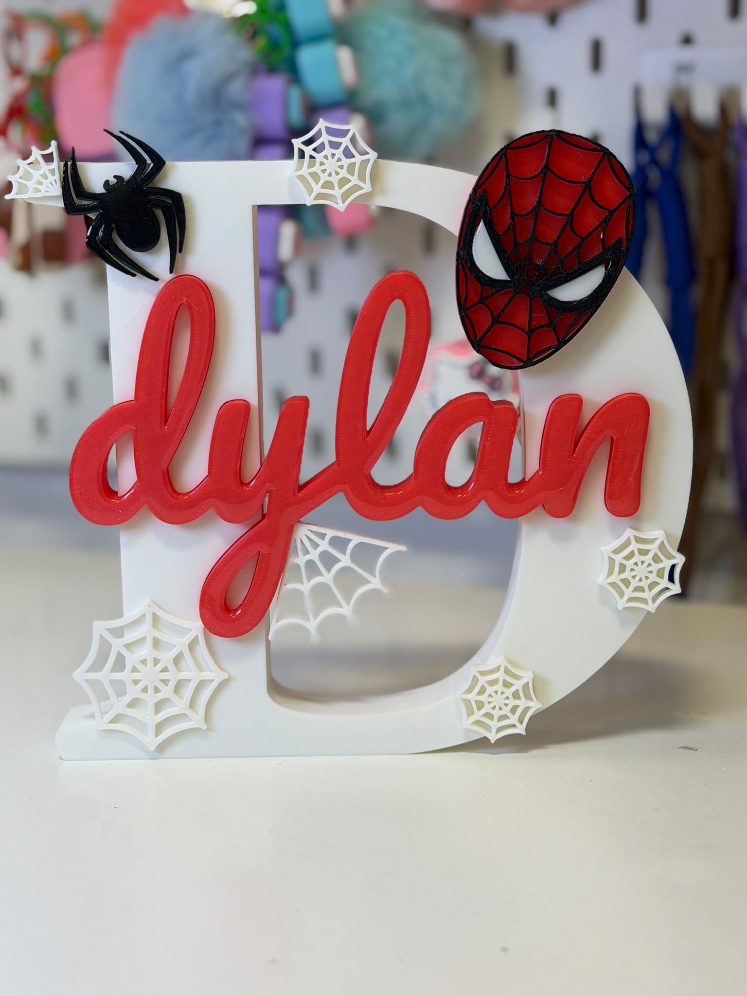 Letra D blanca con el nombre dylan en caligrafía roja, máscara arácnida roja, araña negra y telarañas blancas en relieve