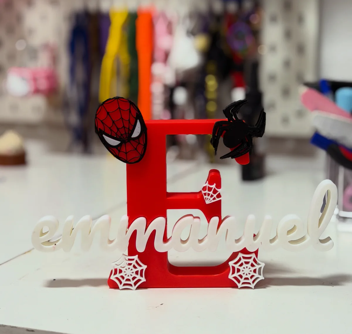 Letra E roja decorada con telaraña y personaje arácnido, con el nombre emmanuel en letras cursivas blancas debajo